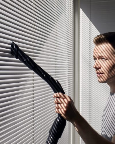 Man cleaning dust from window blind by vacuum cleaner